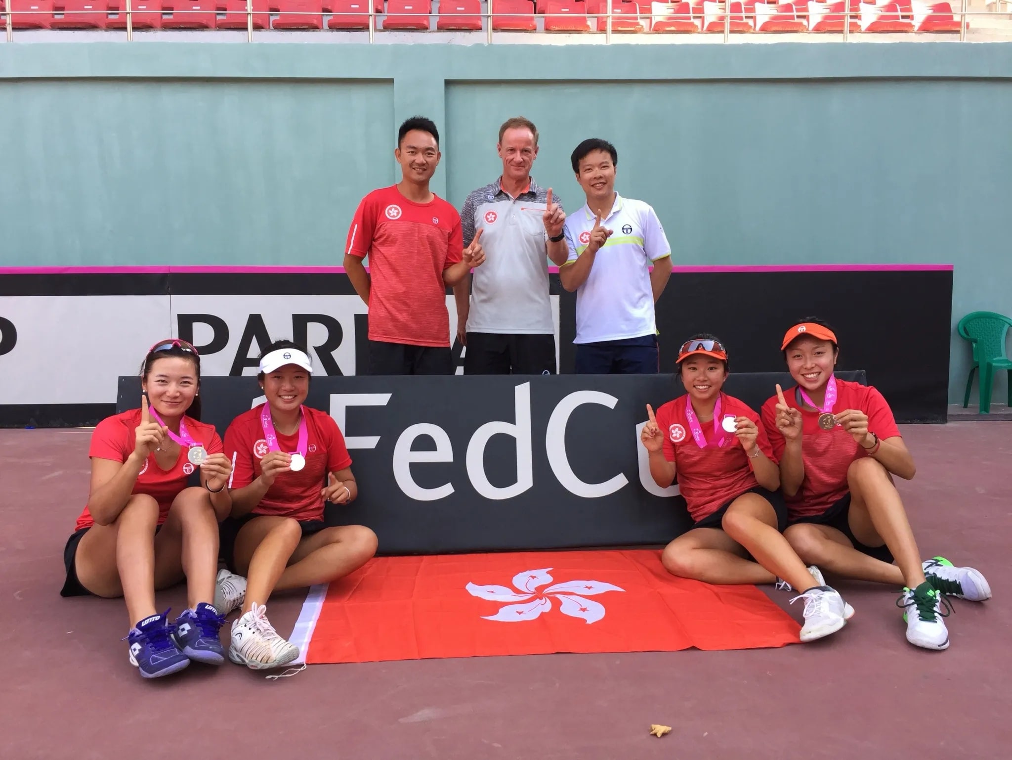 Hong Kong players with their medals, seated around the Hong Kong flag in front of a Fed Cup board, with Jeff King and two coaches standing behind.