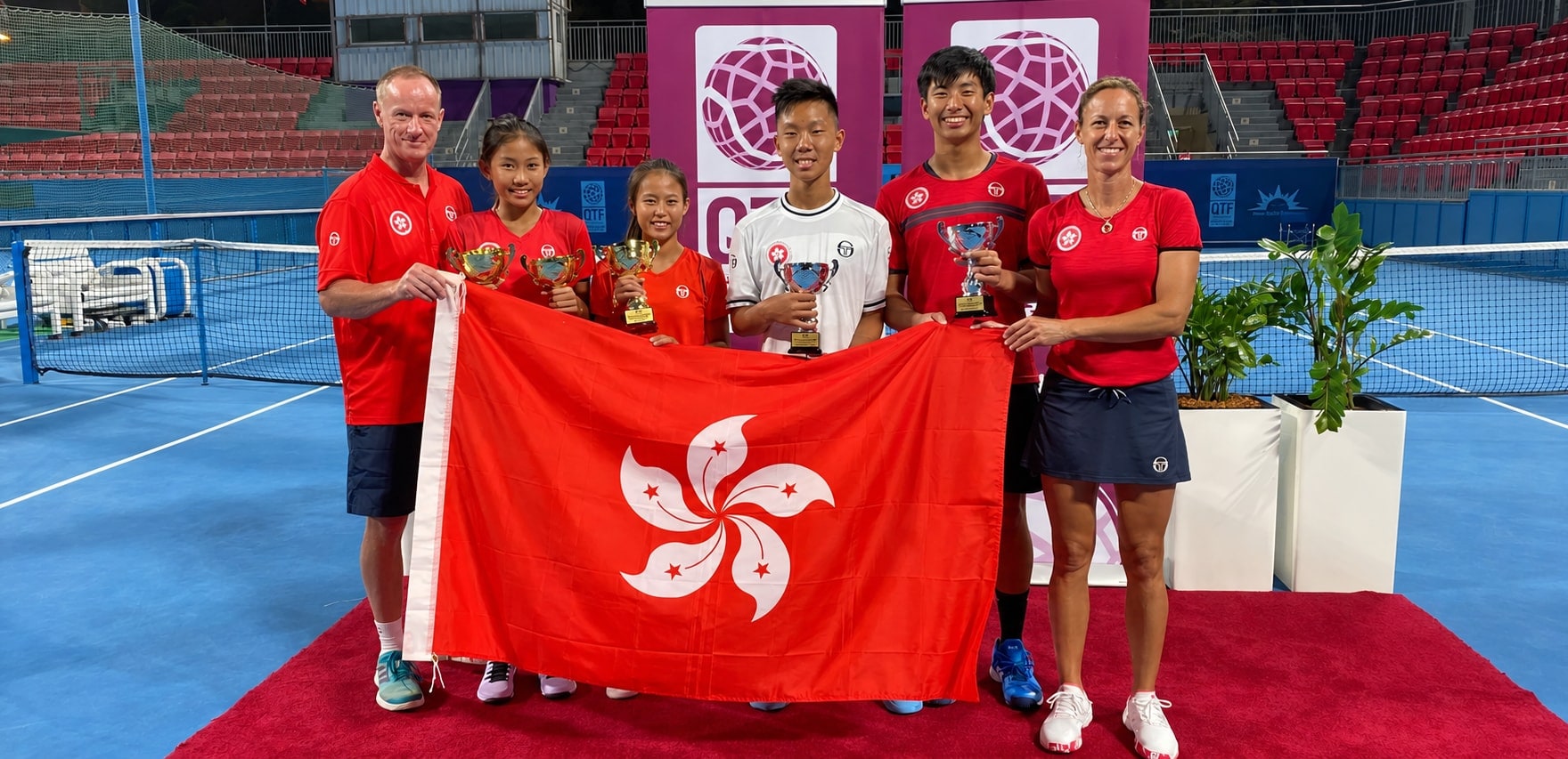 Jeff King and a fellow coach with four Hong Kong juniors holding trophies and the Hong Kong flag at a Qatar Tennis Federation event.